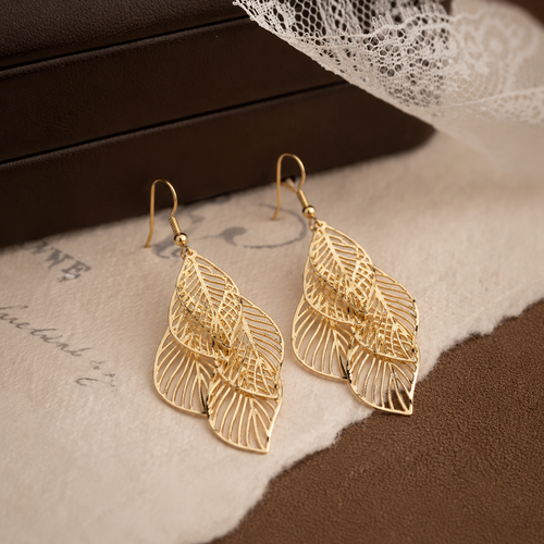 A pair of Elnovaire gold-toned drop earrings with an intricate openwork leaf design, displayed on a textured beige surface with a torn-paper effect over a brown background.