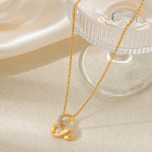 A Elnovaire gold chain necklace with an interlocking knot pendant, one loop set with small clear stones, displayed on a cream satin fabric next to a clear glass pedestal.