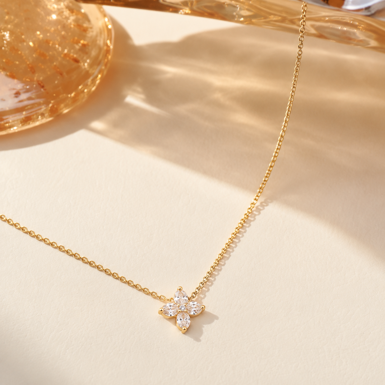 A Elnovaire delicate gold chain necklace with a small flower-shaped pendant set with clear stones, displayed on a warm beige surface with soft shadows.
