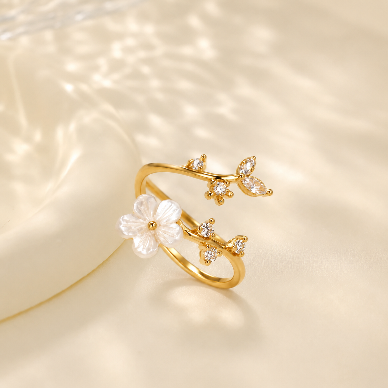 A Elnovaire delicate gold ring designed like a branch with a white flower and small clear stones, photographed on a soft cream surface with dappled light shadows.