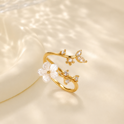 A Elnovaire delicate gold ring designed like a branch with a white flower and small clear stones, photographed on a soft cream surface with dappled light shadows.