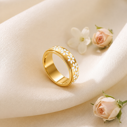 A Elnovaire gold band ring decorated with white floral cutouts, resting on draped cream fabric with small pale pink and white flowers nearby.