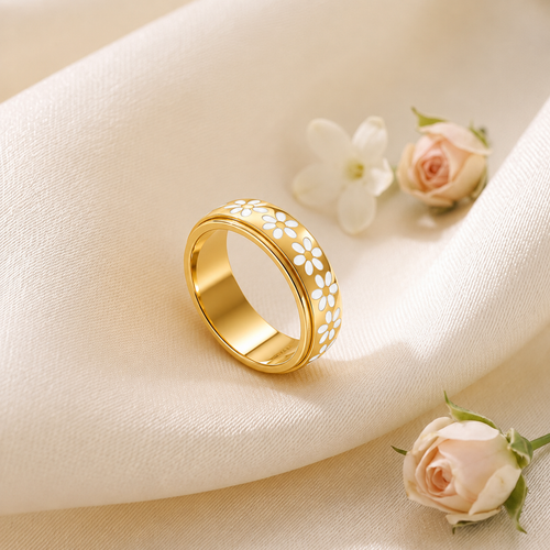 A Elnovaire gold band ring decorated with white floral cutouts, resting on draped cream fabric with small pale pink and white flowers nearby.