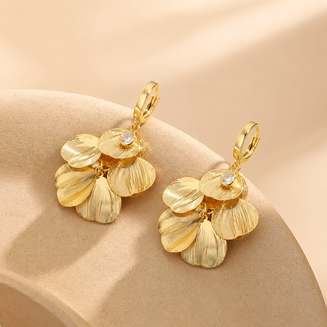 A pair of Elnovaire gold-toned dangle earrings with textured petal-like drops and small clear stones, displayed on a warm beige curved surface.