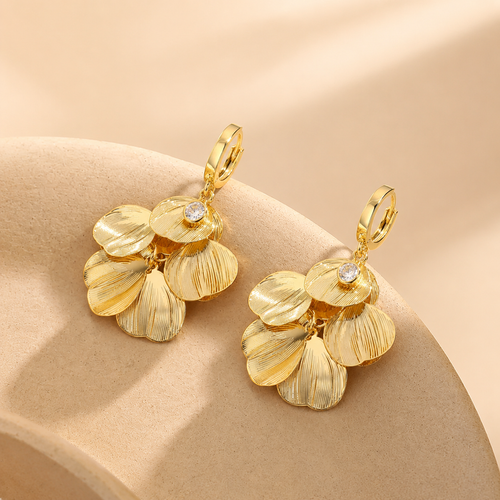 A pair of Elnovaire gold-toned dangle earrings with textured petal-like drops and small clear stones, displayed on a warm beige curved surface.