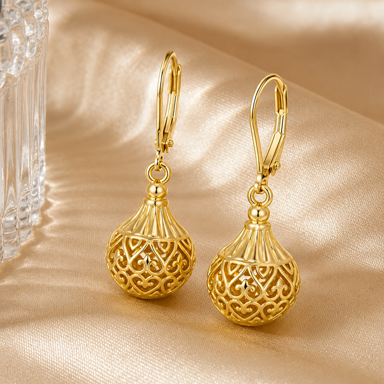 A pair of Elnovaire gold leverback earrings with ornate filigree ball pendants, displayed on a shimmering champagne satin fabric next to a clear glass object.