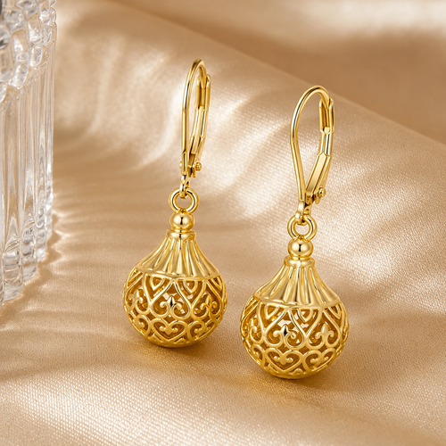 A pair of Elnovaire gold leverback earrings with ornate filigree ball pendants, displayed on a shimmering champagne satin fabric next to a clear glass object.