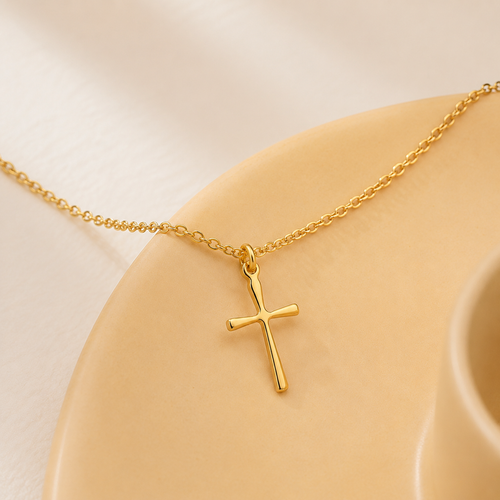 A delicate Elnovaire gold cross pendant necklace resting on a beige ceramic dish against a soft cream background.