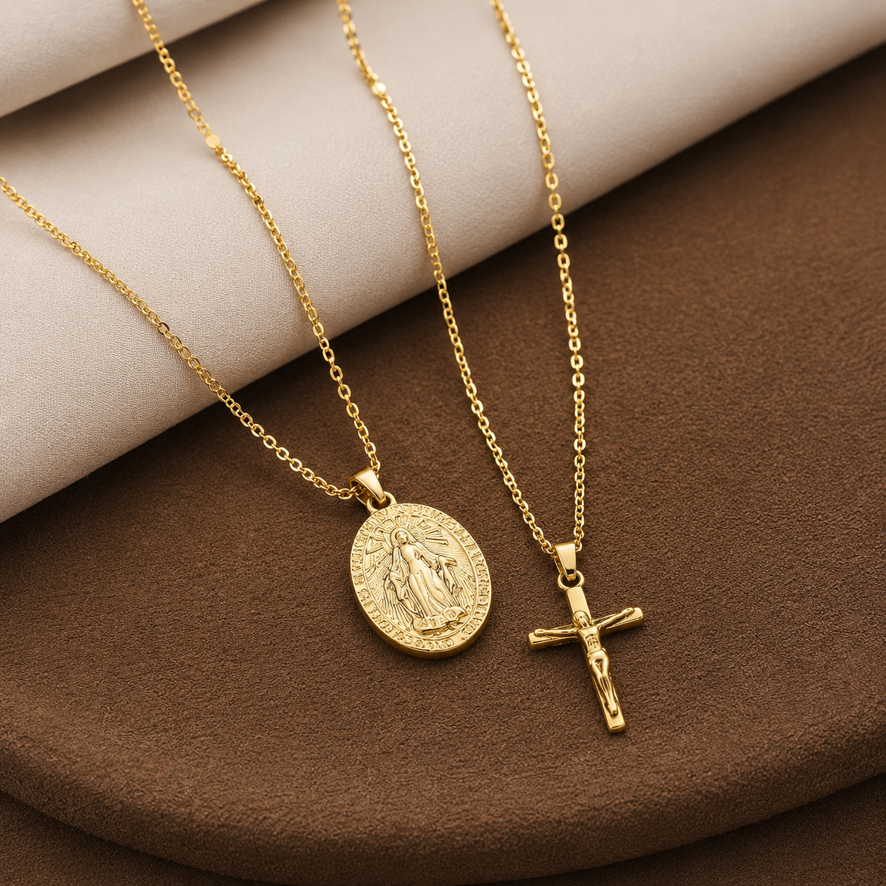 Two Elnovaire gold-tone religious necklaces displayed on a brown and beige fabric surface. One necklace features an oval pendant with a raised figure, and the other features a crucifix pendant.