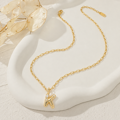 A Elnovaire gold chain necklace with a stylized letter 'K' pendant adorned with small clear stones, displayed on a white sculptural tray with dried botanical elements.
