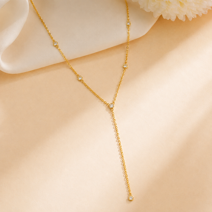 A Elnovaire delicate gold Y-necklace with small clear gemstone accents at each junction and a long drape, displayed on a soft beige fabric background with a white flower prop.